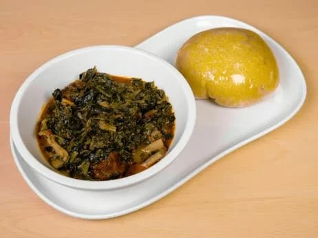 A white oval plate with a bowl of cooked leafy greens and a yellow bread roll on a wooden surface.
