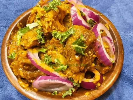 Bowl of Indian curry topped with chopped cilantro, sliced red onions, and green chili peppers, served in a wooden bowl on a blue surface.