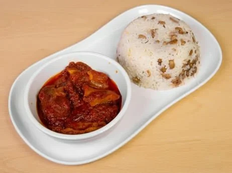 A white divided plate with one side containing a serving of curry with chunks of meat in a red sauce and the other side holding a ball of rice with visible grains and some mixed-in ingredients.