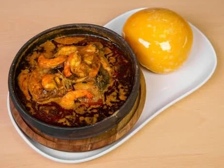Bowl of shrimp and vegetable curry with a yellow ball-shaped bread or pudding on a white plate.