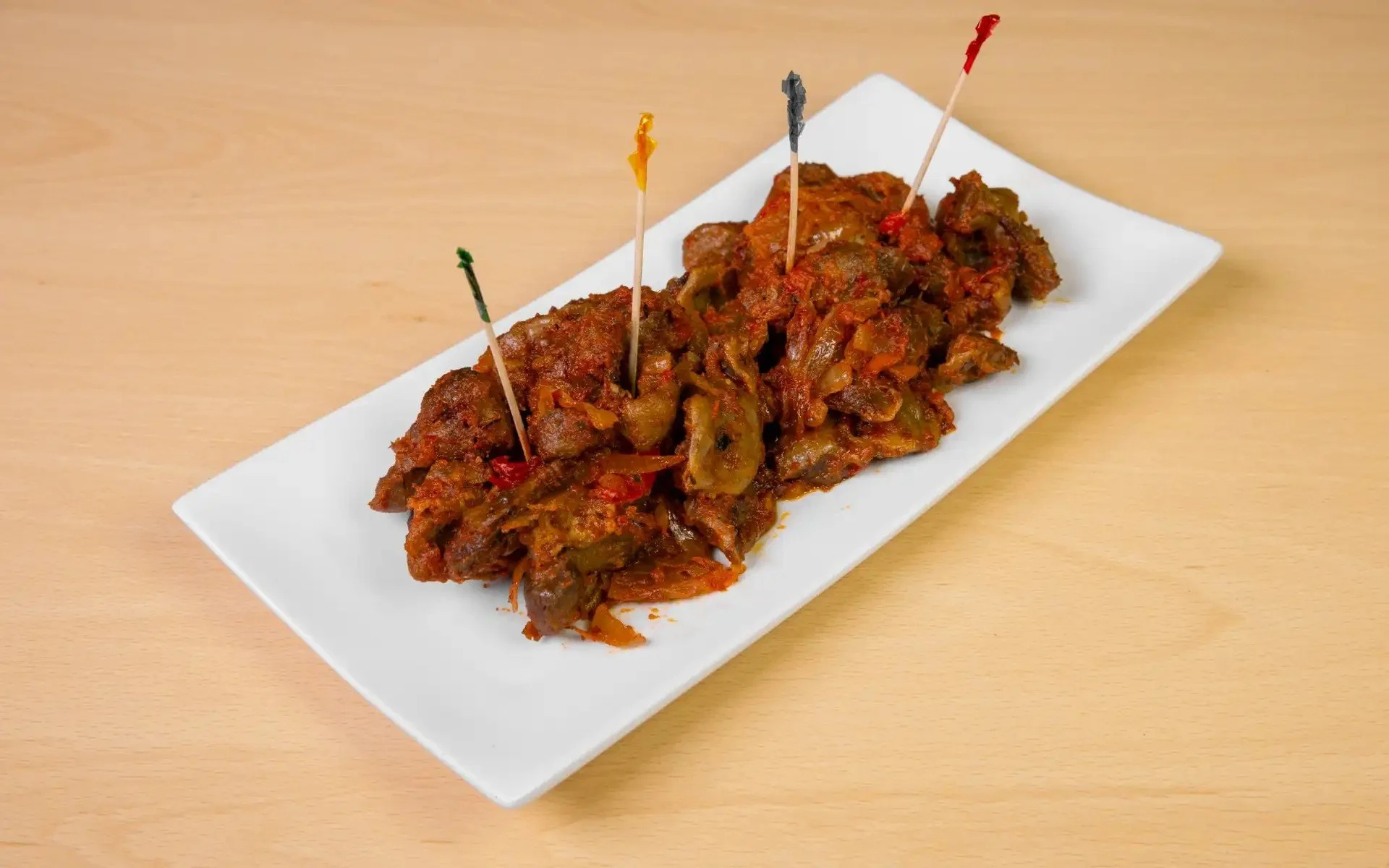 Plate of spicy cooked meat and vegetables garnished with small colorful flags on toothpicks, on a white rectangular dish