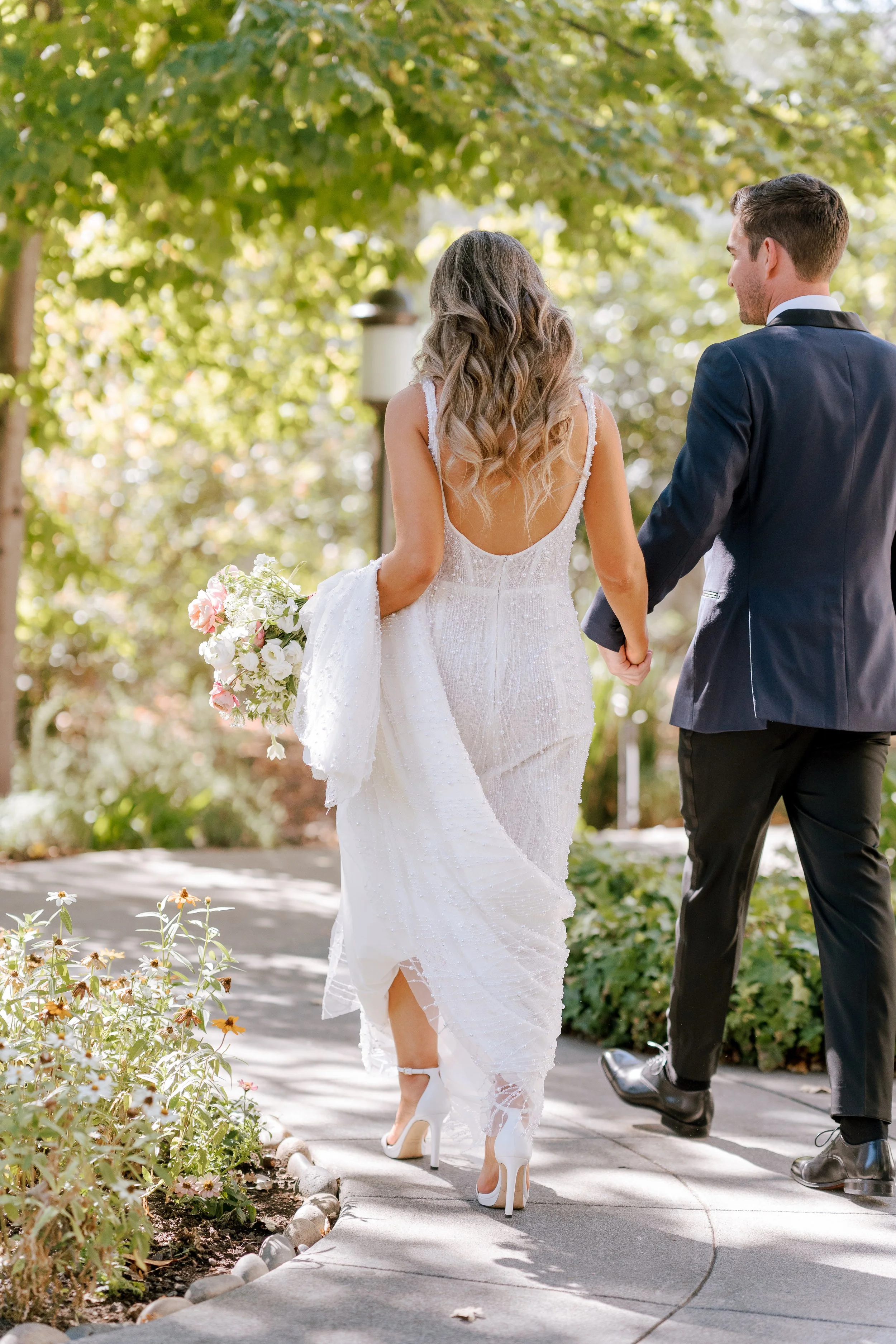 A bride and groom holding hands walking outdoors, with the bride in a white wedding dress and high heels, and the groom in a dark suit.