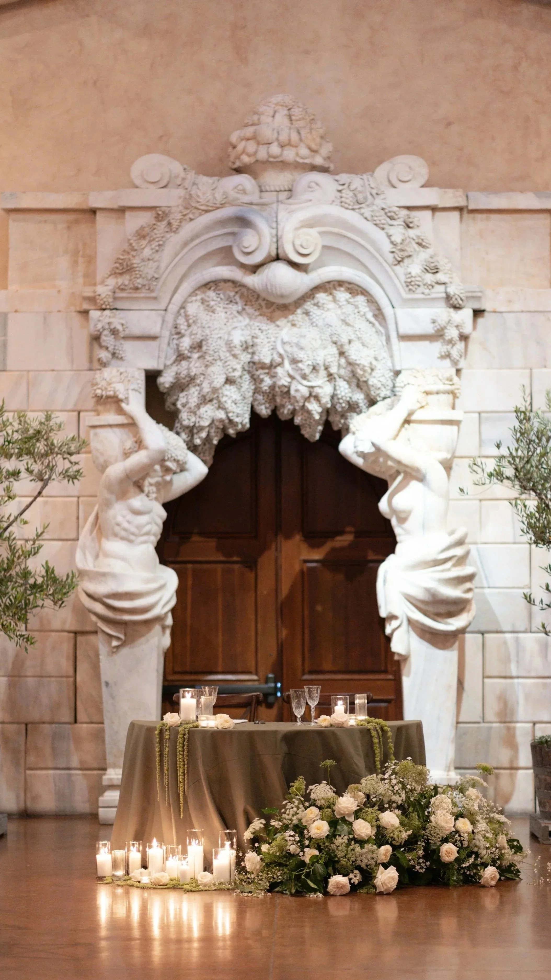 Wedding or formal event setup with a large white marble arch decorated with sculptures and floral arrangements, candles, and a table with drinks and flowers in front.