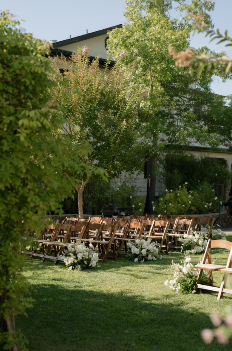 Outdoor wedding ceremony setup with wooden chairs arranged on green grass, decorated with white floral arrangements, surrounded by trees and greenery.