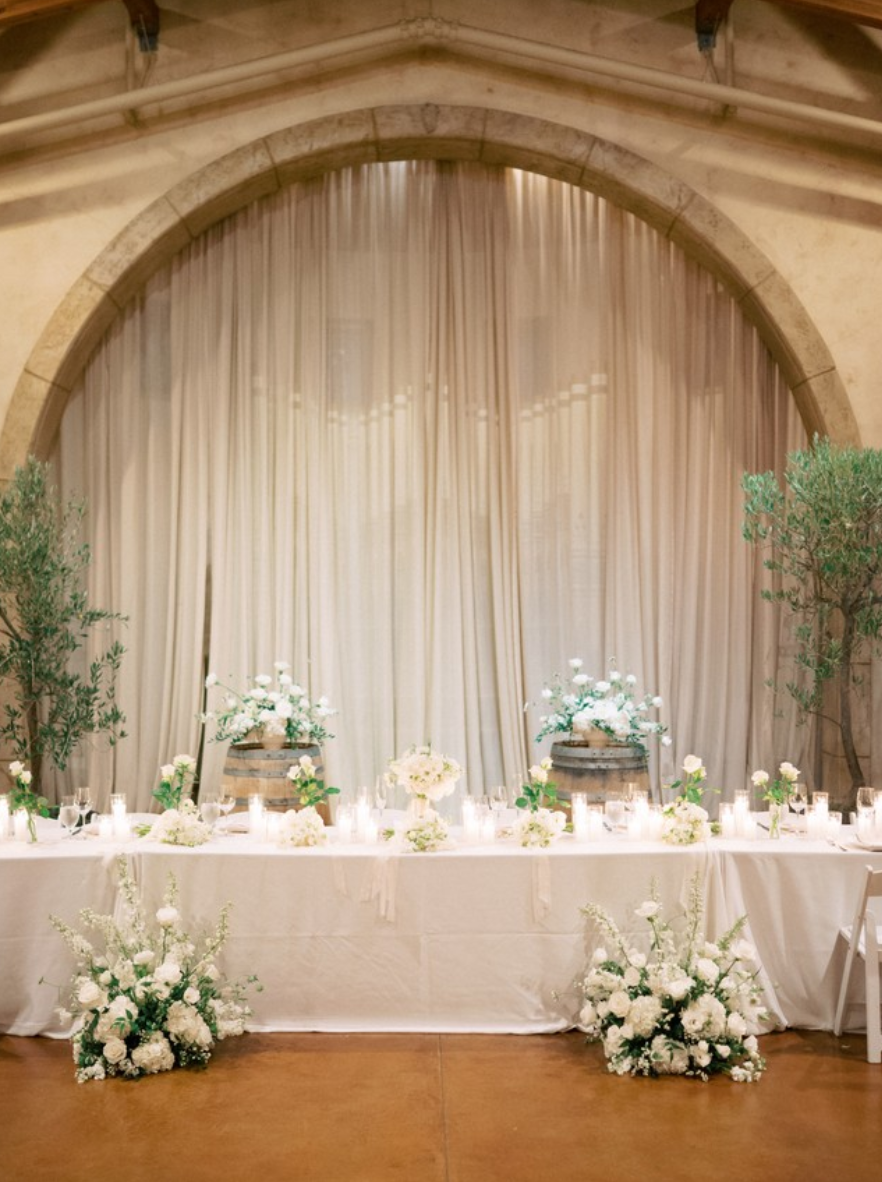 Elegant wedding reception table decorated with white flowers, candles, and greenery, set against a backdrop of cream curtains and an arched stone wall.