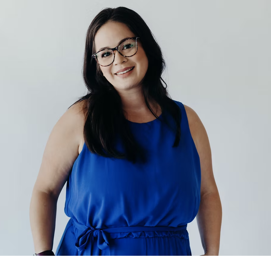 A smiling woman with glasses and long dark hair wearing a blue sleeveless dress standing against a plain light background.