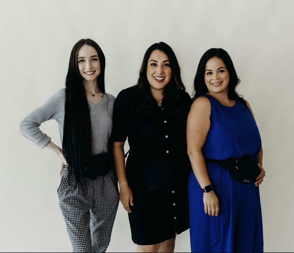 Three women smiling and standing together against a plain wall, dressed in stylish, casual clothing.