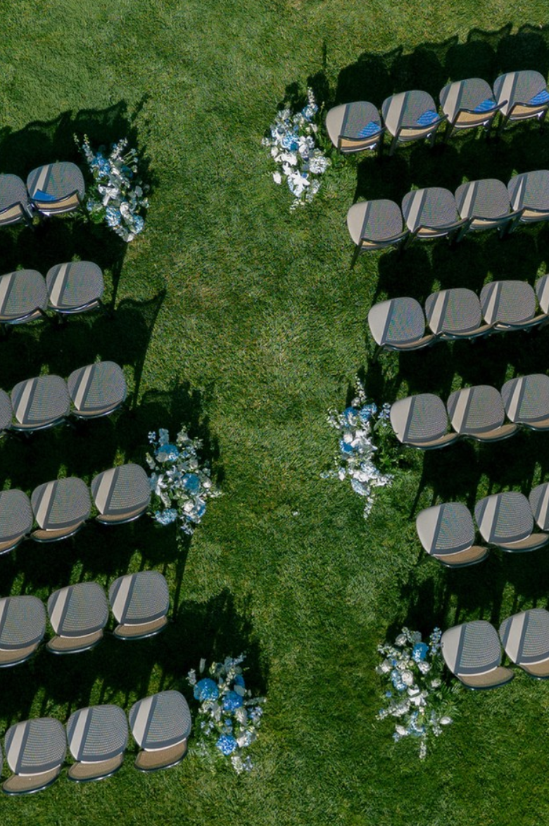 Empty chairs arranged in rows at an outdoor wedding ceremony with floral arrangements along the aisle on a grassy lawn.