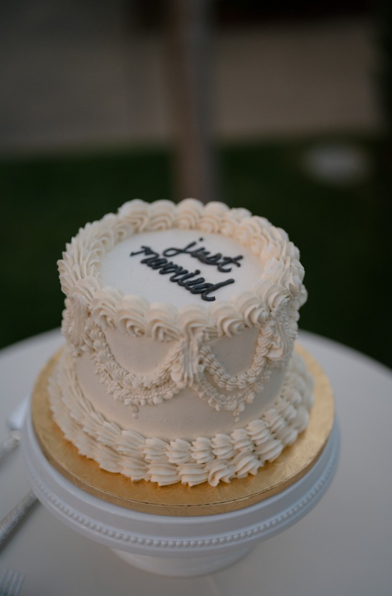 A white wedding cake with piped cream decorations and the words 'Just Married' written on top in black icing.