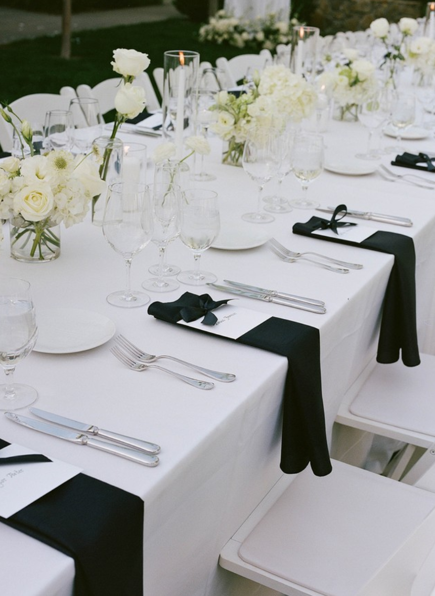 Elegant outdoor dining table decorated with white flowers, candles, glassware, and black napkins with ties, set for a formal event.