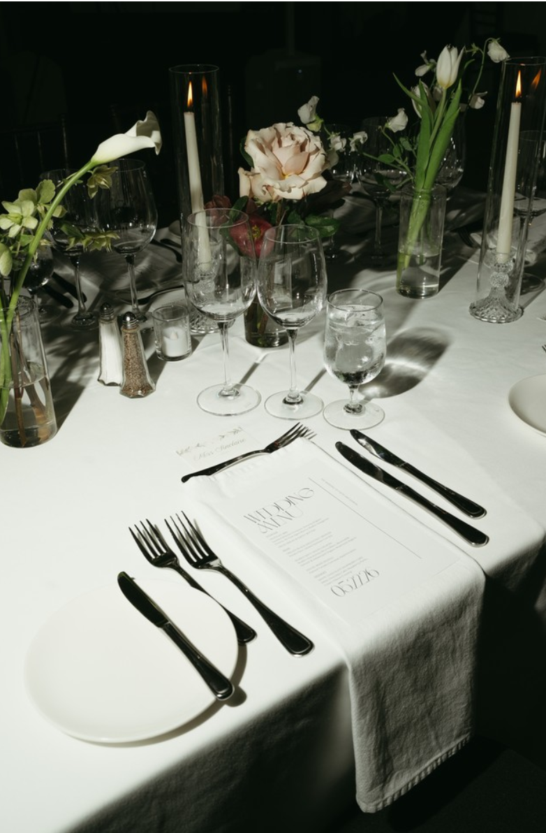 Elegant dining table setup with white tablecloth, wine glasses, water glasses, floral centerpieces, taper candles, and silverware, ready for a formal meal.