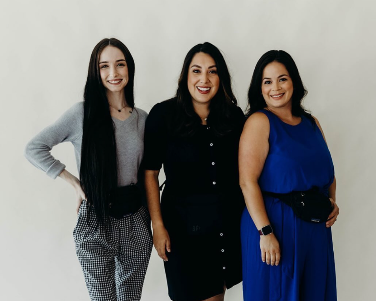 Three women standing together, smiling, against a plain light-colored background.