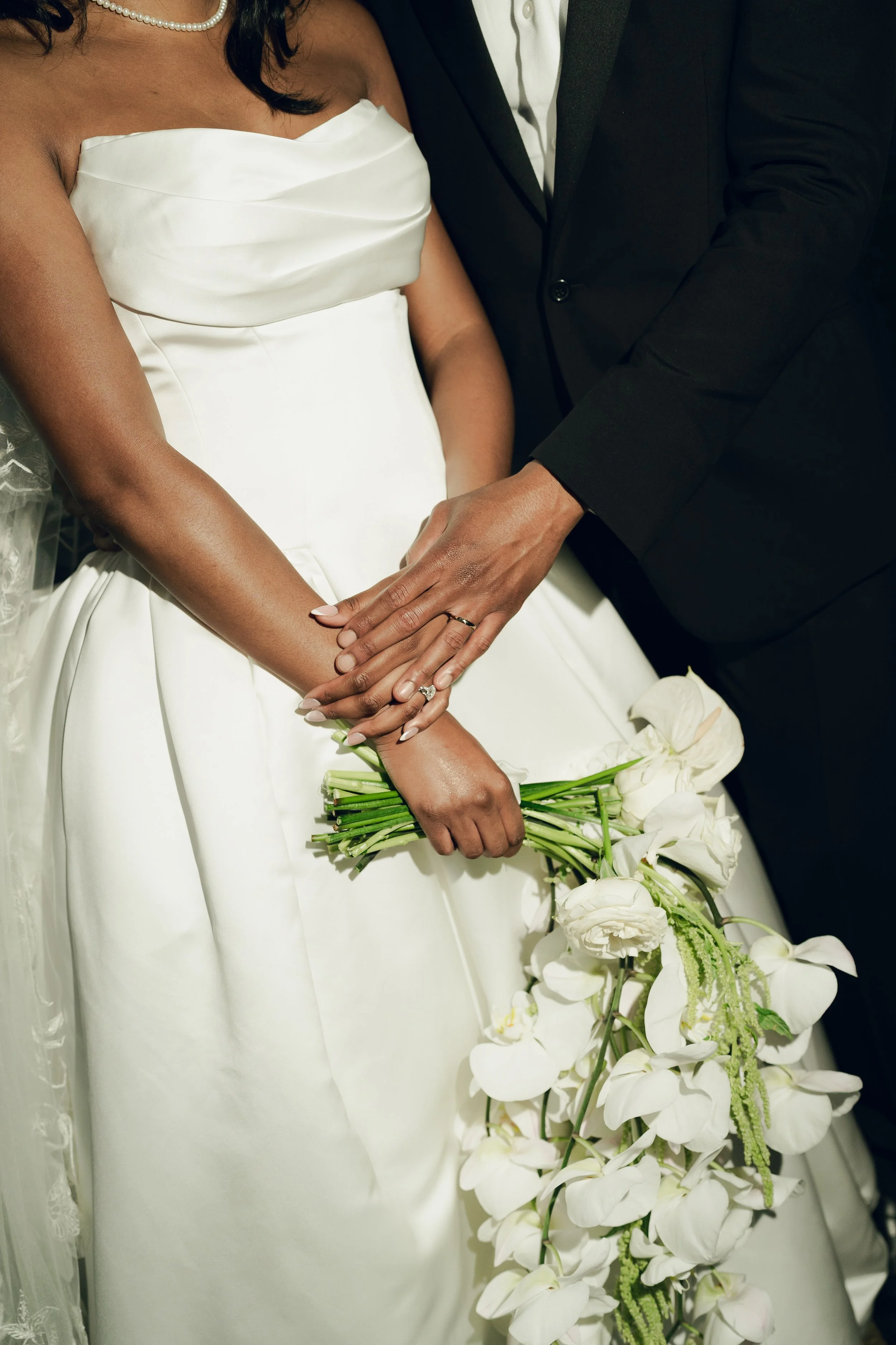 Close-up of a bride and groom holding hands, with the bride wearing a white satin wedding gown and veil, and the groom in a black suit. The bride has a diamond ring on her left hand, and they are holding a cascading bouquet of white flowers.