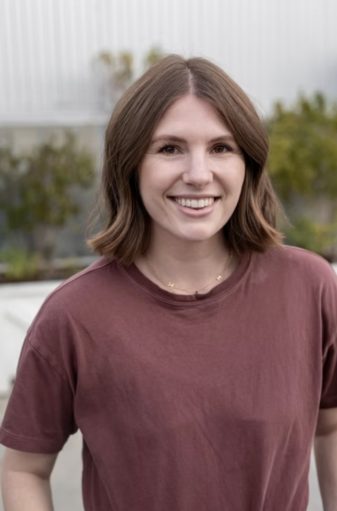 A young woman with shoulder-length brown hair smiling outdoors, wearing a maroon T-shirt and a small gold necklace, with blurred greenery and a structure in the background.