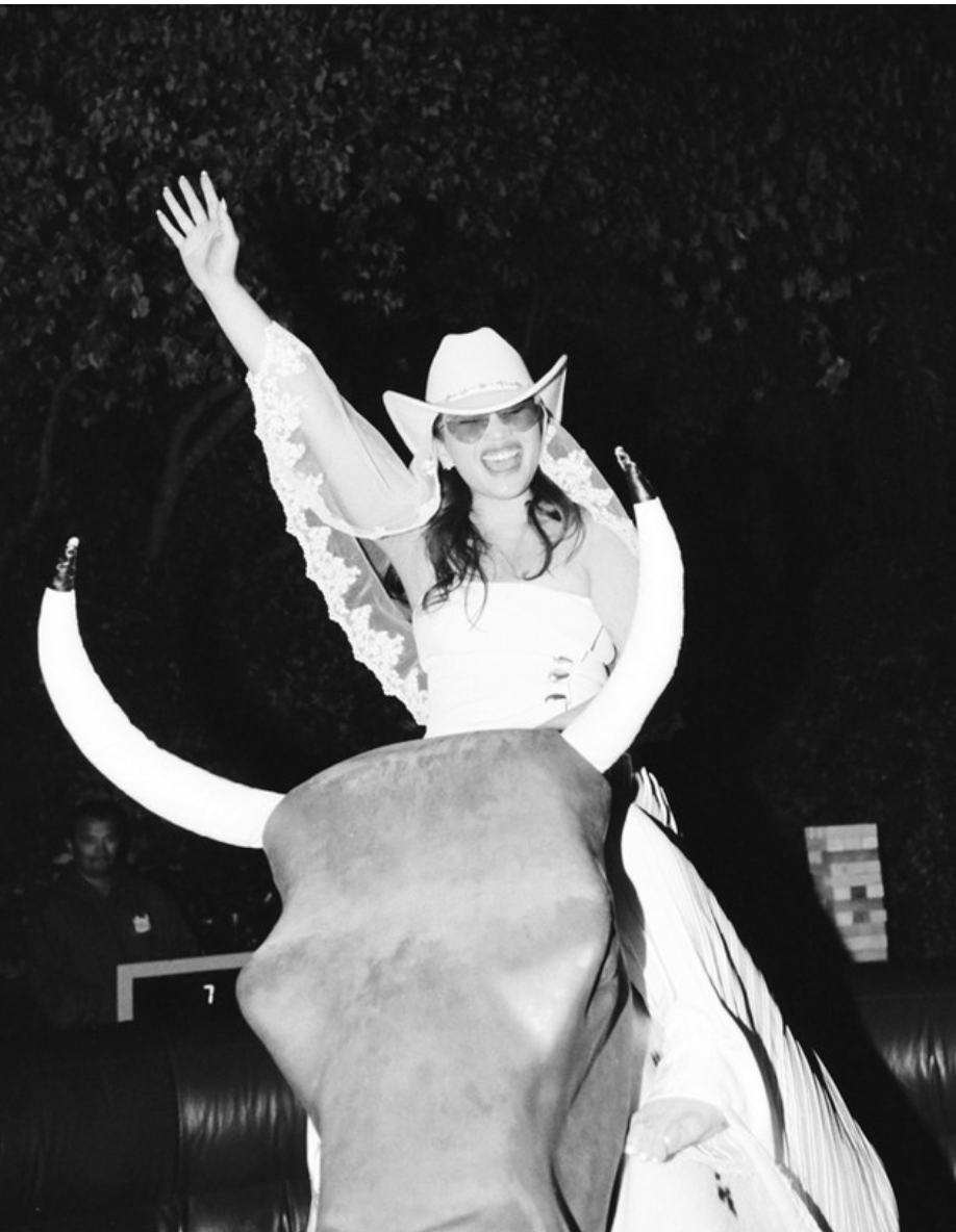 A woman wearing a cowboy hat and sunglasses, smiling and raising her hand, appears to be riding a mechanical bull during a nighttime event outdoors.