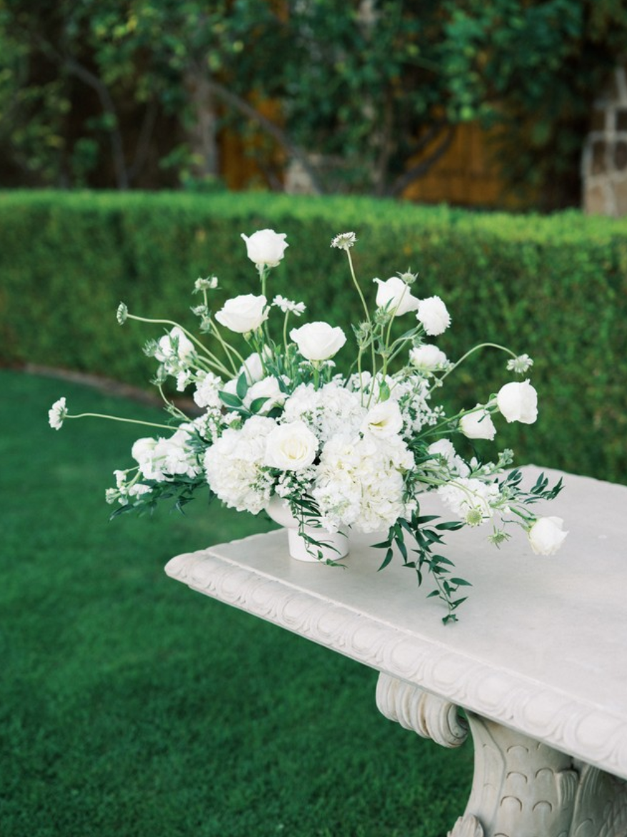 White floral arrangement in a vase on a white ornate table outdoors, with green bushes and trees in the background.