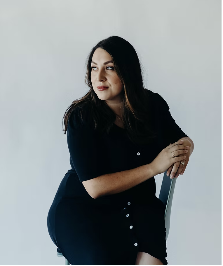 A woman with dark hair, wearing a black dress, sitting on a chair against a plain white wall, looking to the side with a thoughtful expression.