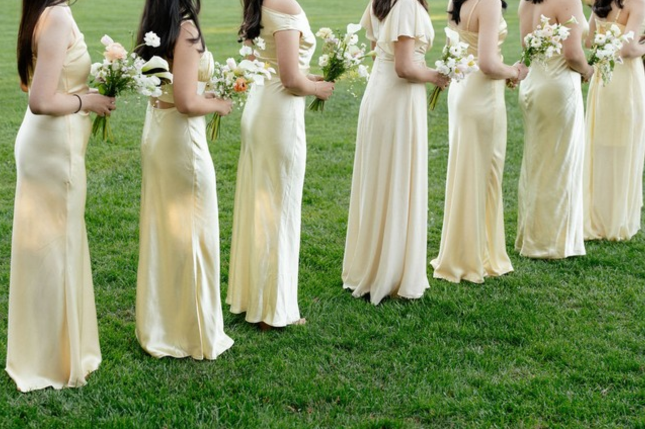 A line of bridesmaids in yellow dresses holding bouquets standing on grass at a wedding.