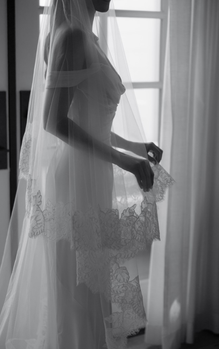 Silhouette of a bride in wedding dress with veil, standing near a window, holding lace fabric, in black and white.
