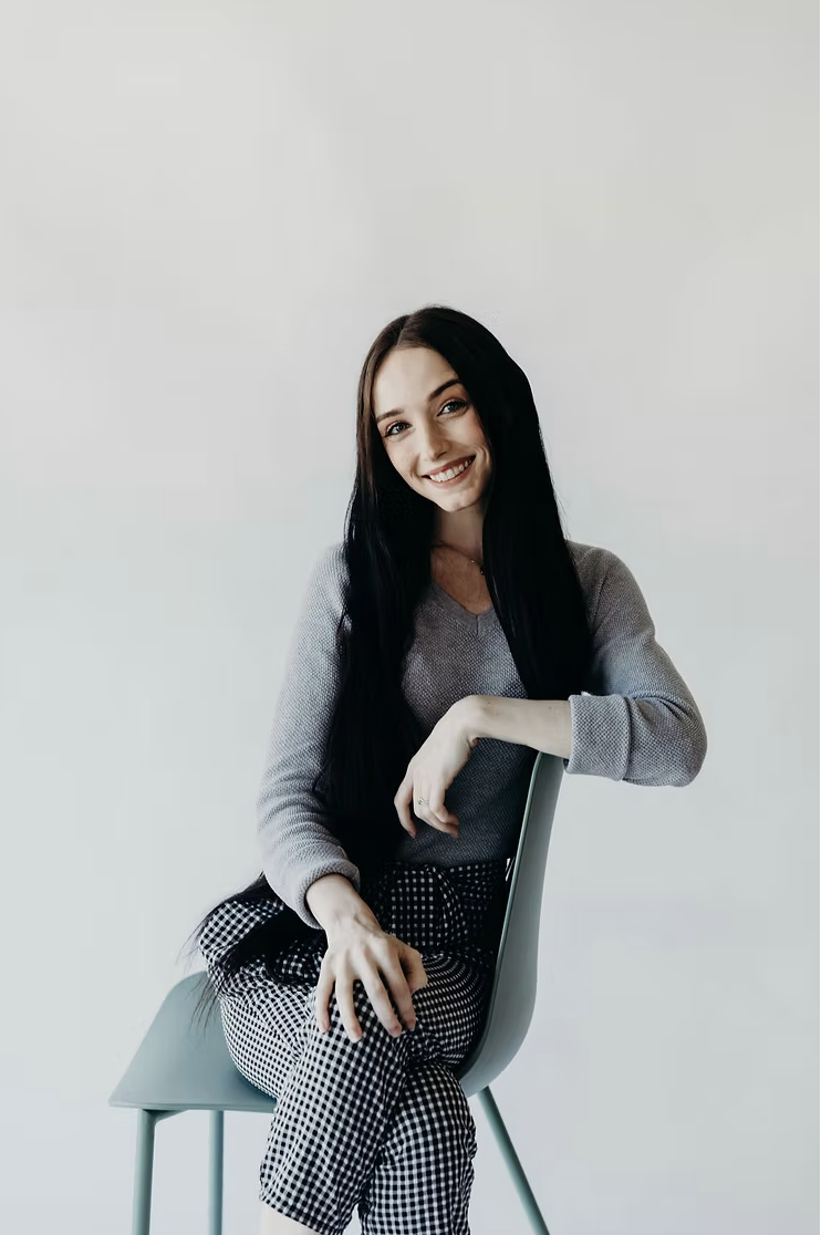 A young woman with long dark hair is sitting on a chair, smiling and leaning slightly on the backrest, against a plain white background. She is wearing a gray long-sleeve shirt and black-and-white checkered pants.