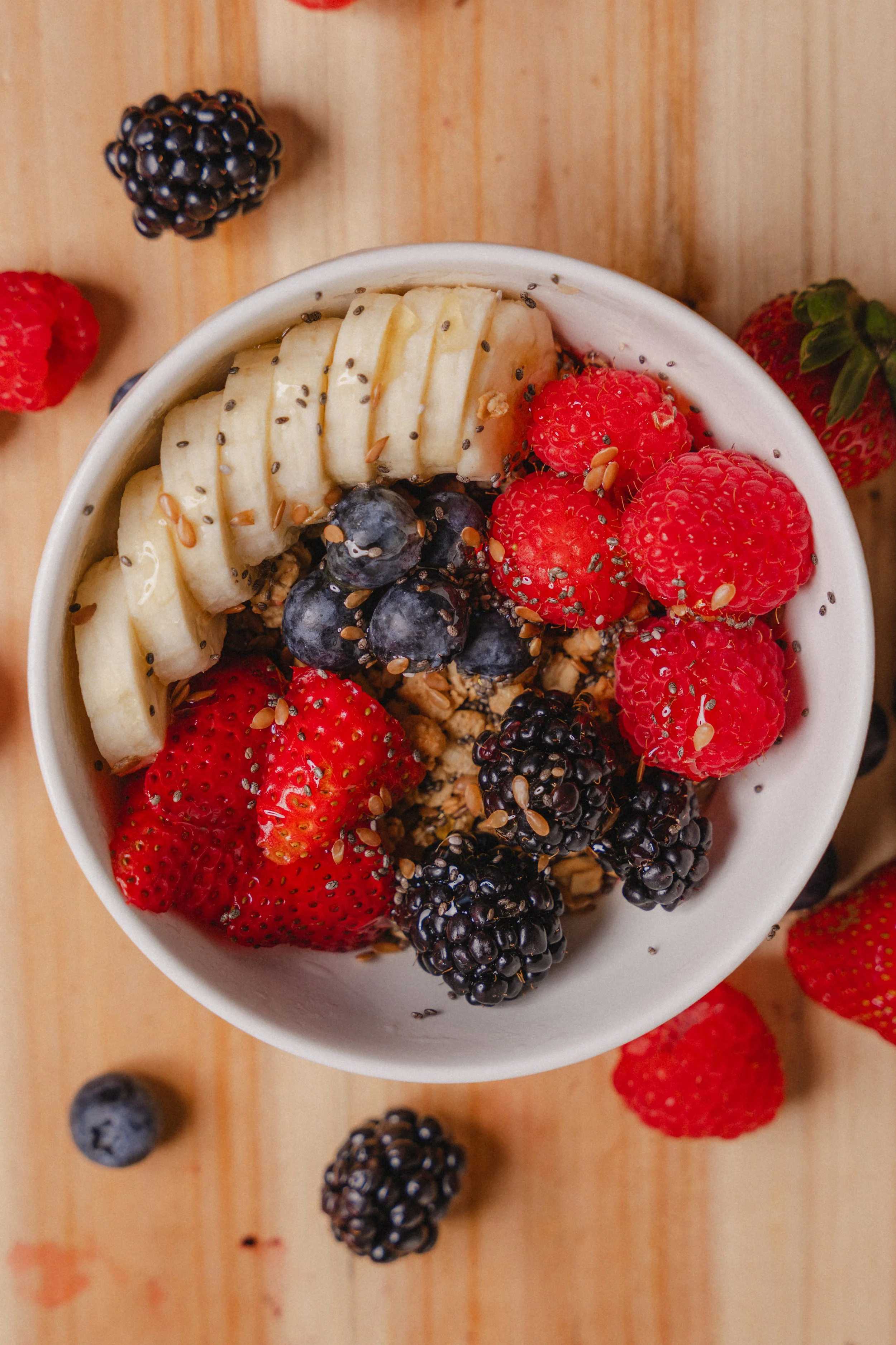 A bowl filled with sliced bananas, strawberries, blueberries, blackberries, and topped with chia seeds and flax seeds on a wooden surface.