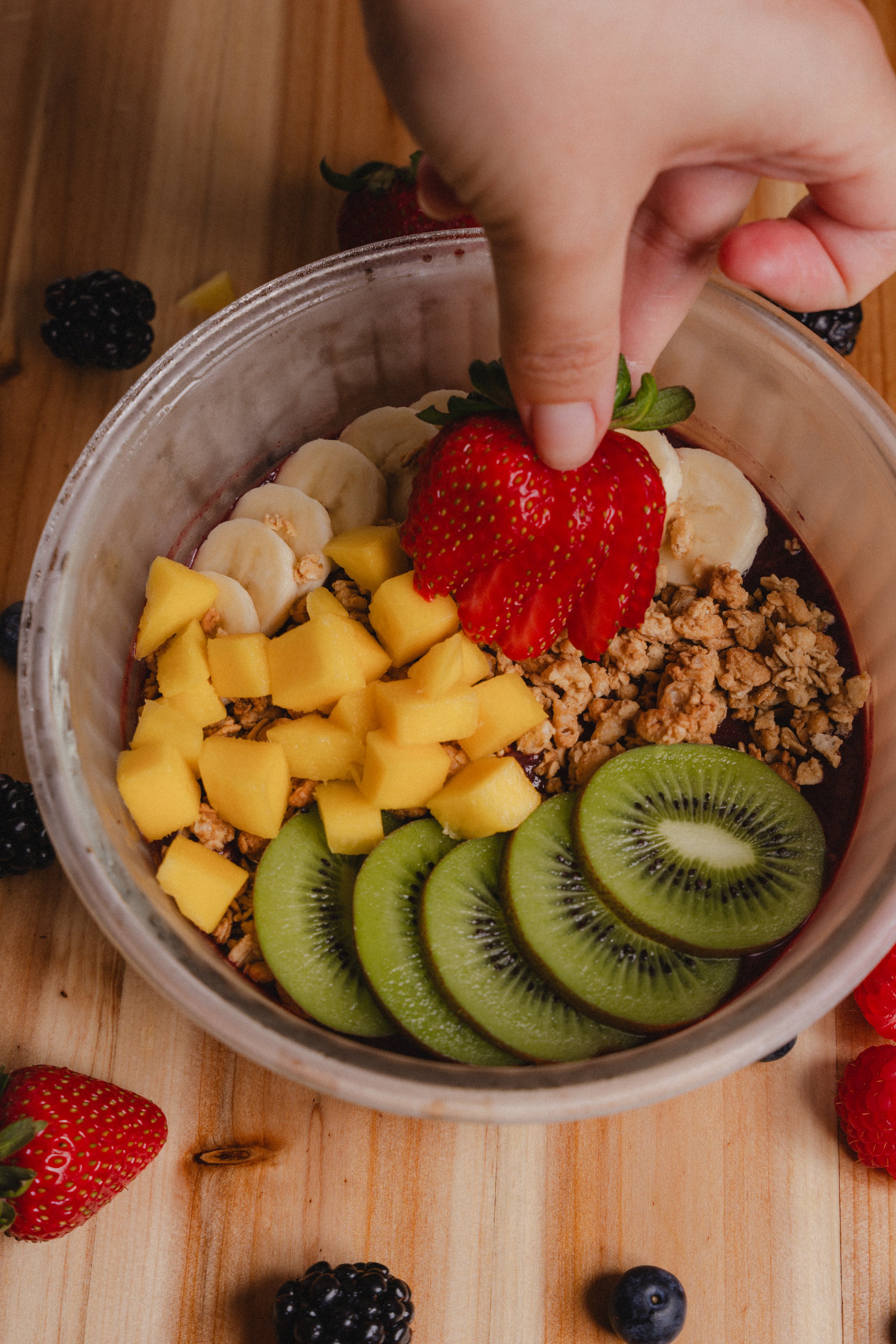 A clear plastic bowl filled with sliced strawberries, bananas, mango, kiwi, blackberries, blueberries, and granola on a wooden surface.