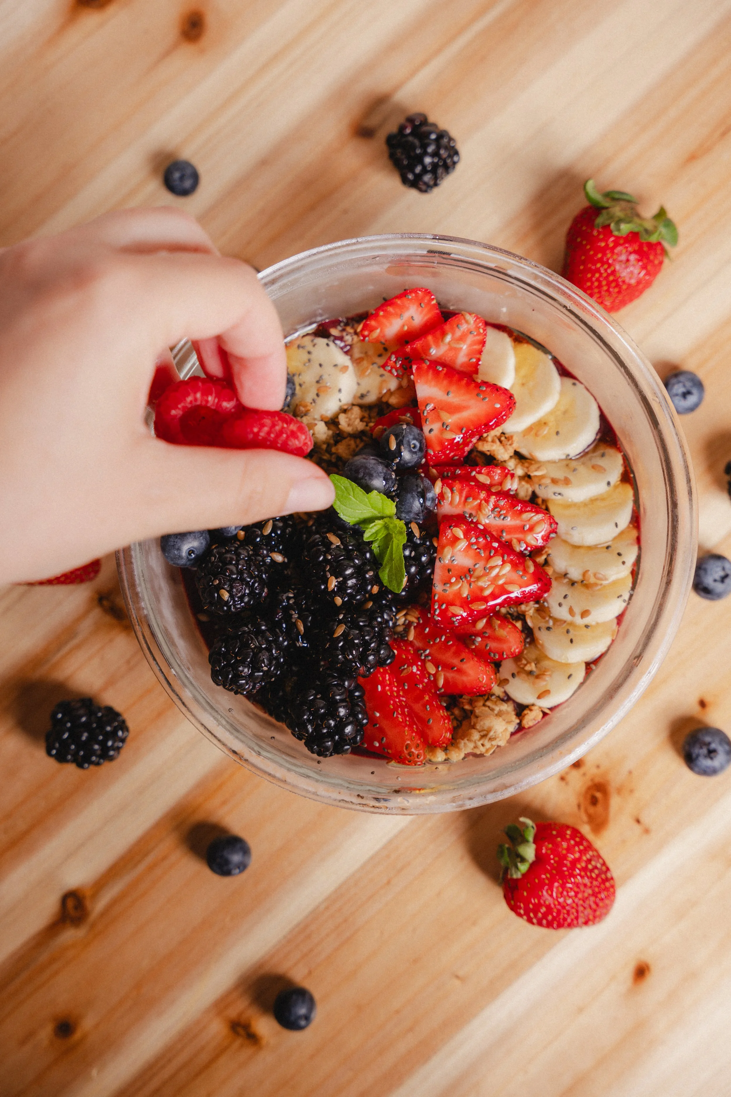 A glass jar filled with sliced strawberries, bananas, blackberries, blueberries, granola, and topped with seeds and a sprig of mint. A hand is placing a raspberry into the jar, and there are loose berries on a wooden surface around it.