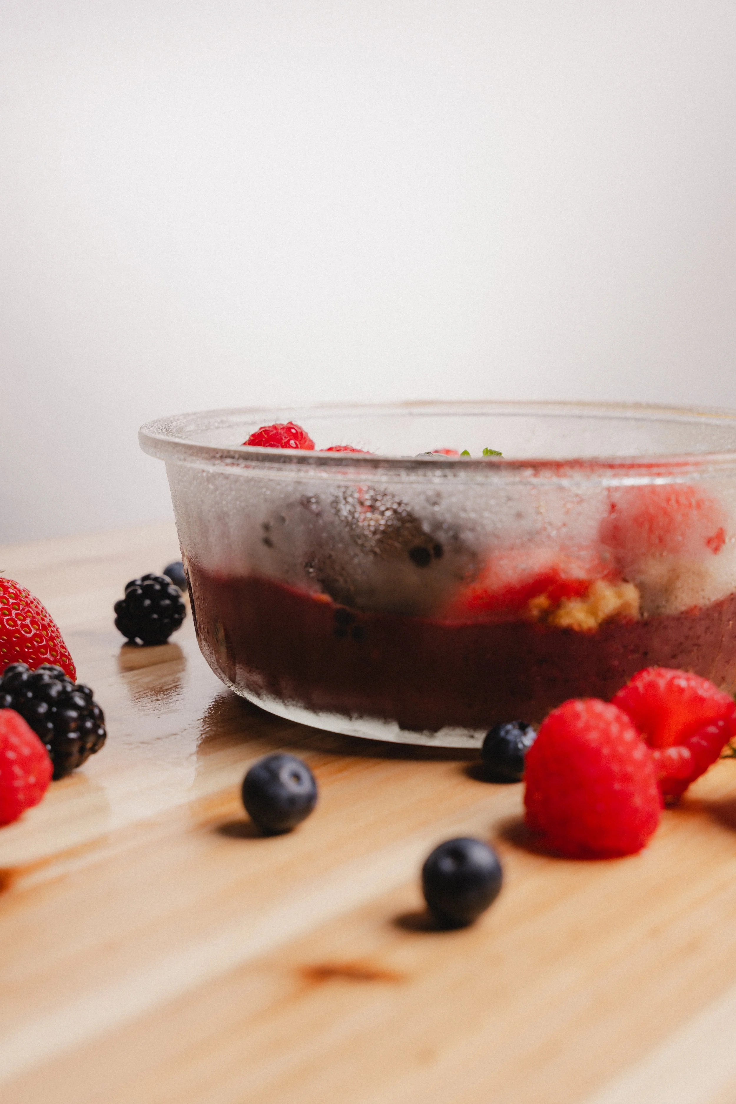 Close-up of a glass bowl with mixed berries and dessert, surrounded by scattered berries on a wooden surface.
