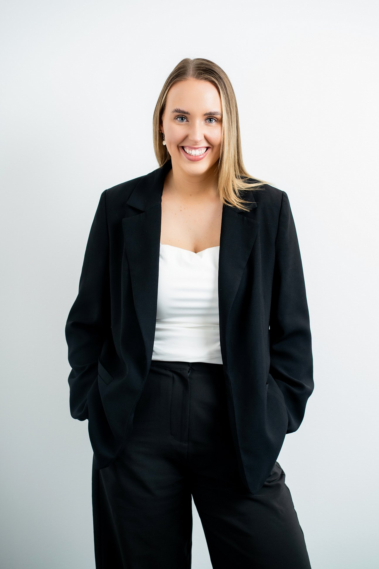 A woman with shoulder-length blonde hair smiling, dressed in a black blazer, white top, and black pants, standing against a plain light gray background.