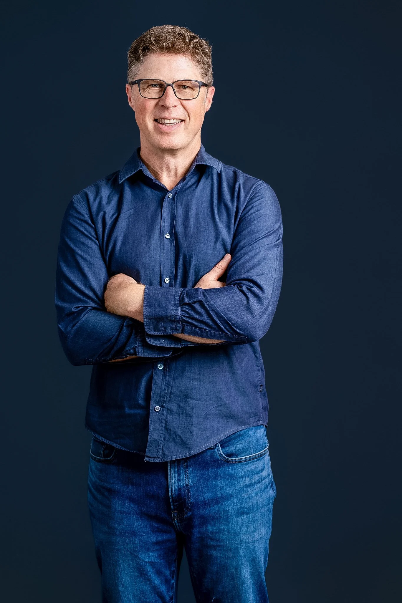 A middle-aged man with glasses, wearing a blue button-up shirt and jeans, standing with arms crossed against a dark background.