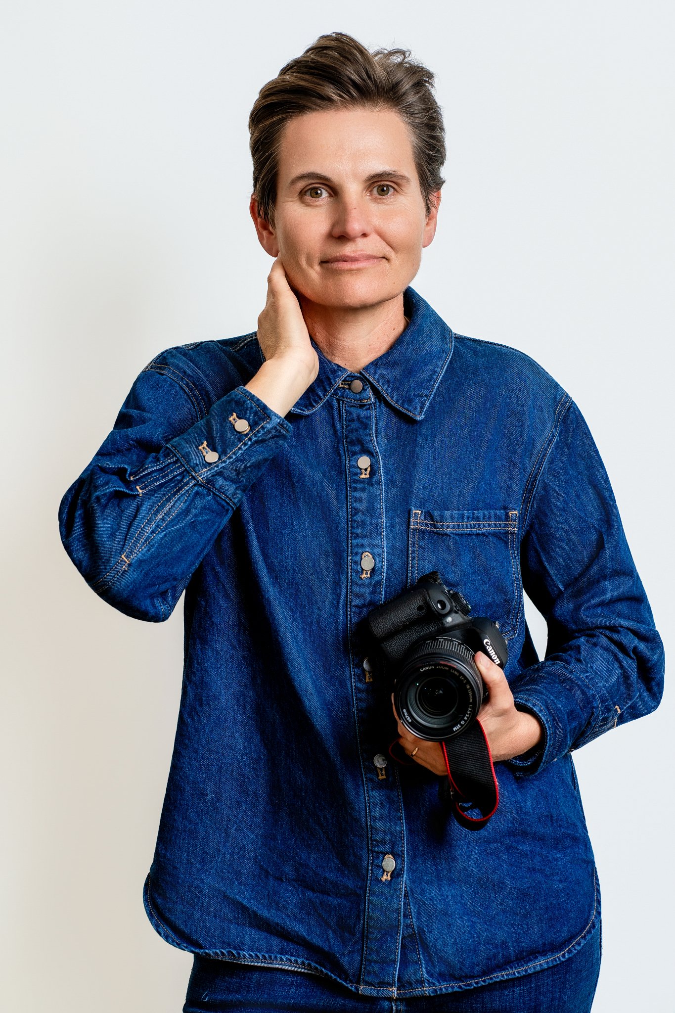 A woman with short brown hair wearing a denim jacket and holding a Canon DSLR camera, standing against a plain light background.