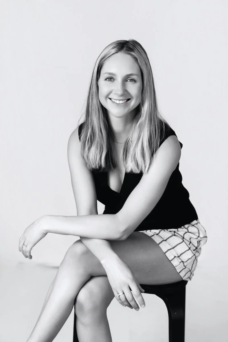 Black and white portrait of a young woman with long hair, smiling, sitting on a stool with her legs crossed, wearing a sleeveless top and checkered shorts.
