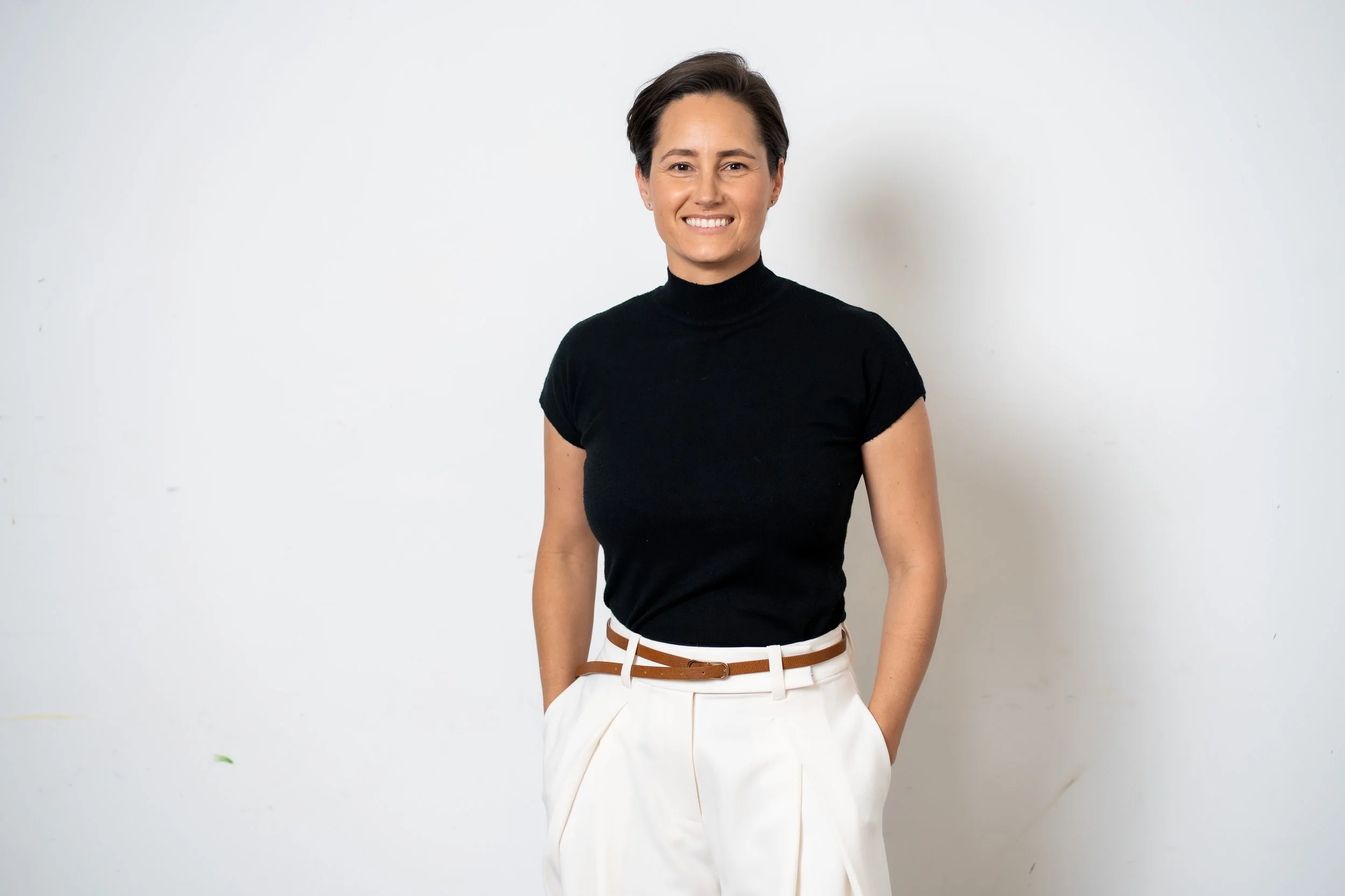A woman with short brown hair wearing a black turtleneck top and white pants, smiling and standing against a plain white background.
