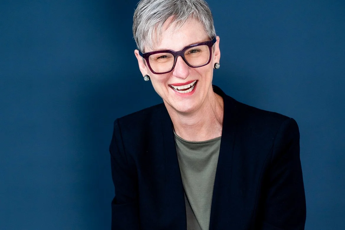 A smiling woman with short gray hair, wearing large black glasses, pearl earrings, a black blazer, and a green shirt, against a solid blue background.