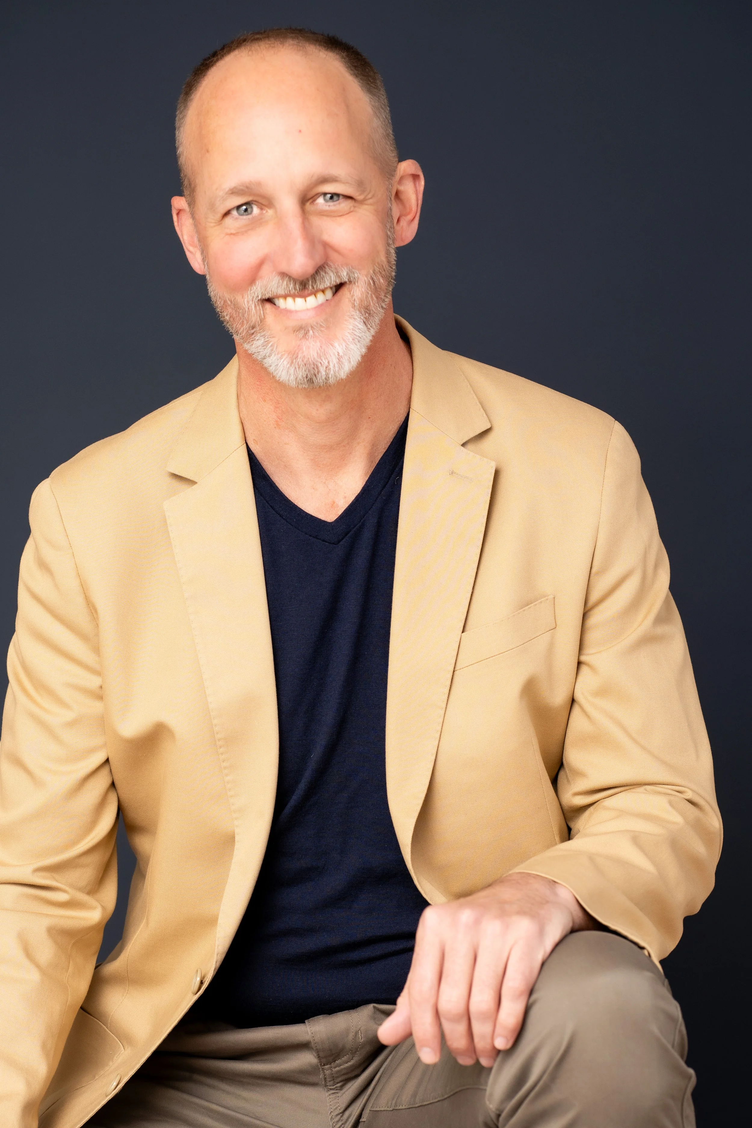 A smiling middle-aged man with a beard and short hair wearing a beige blazer, black shirt, and khaki pants, sitting against a dark background.