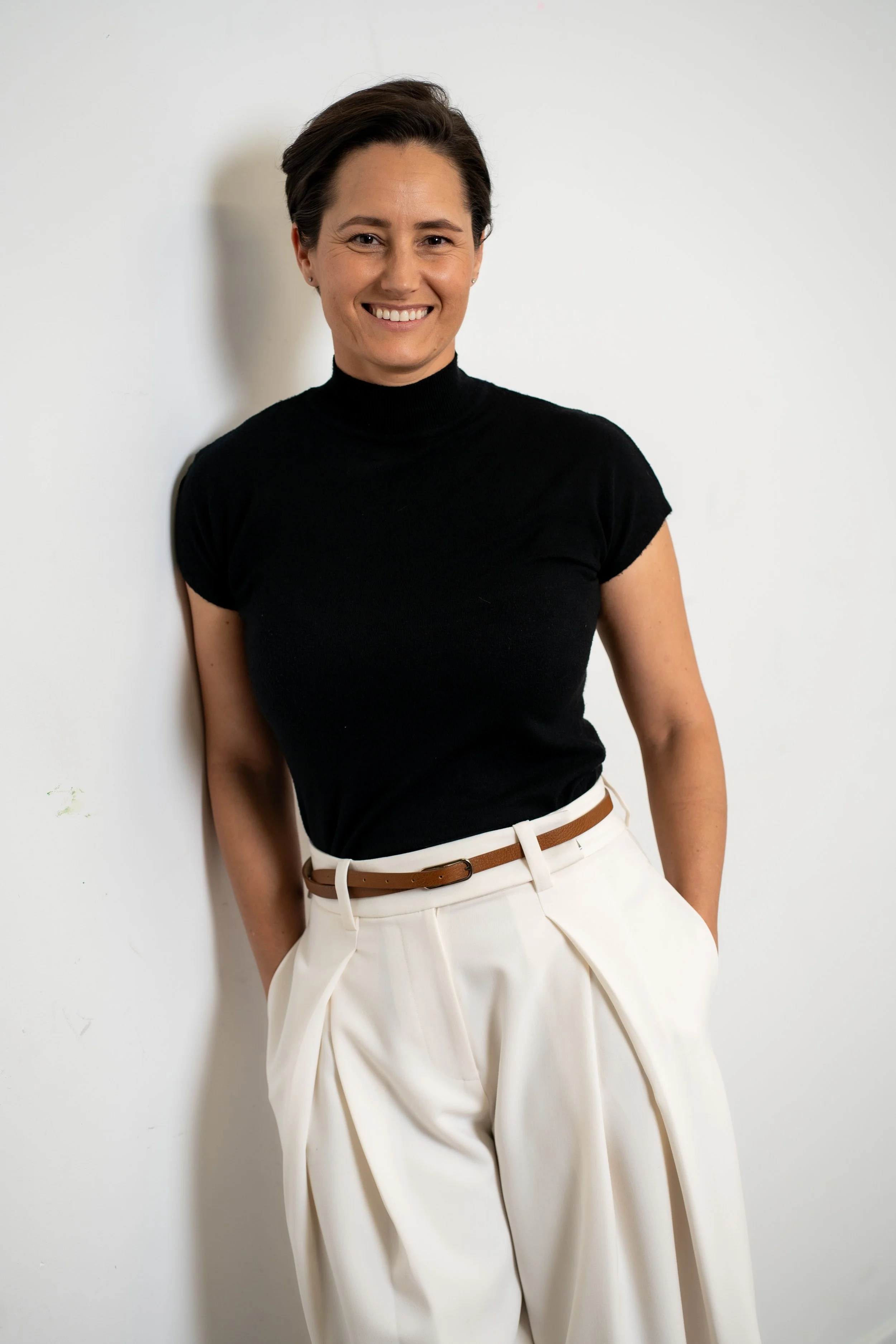A woman with short dark hair wearing a black turtleneck shirt and white wide-leg pants with a brown belt, standing against a white wall, smiling.