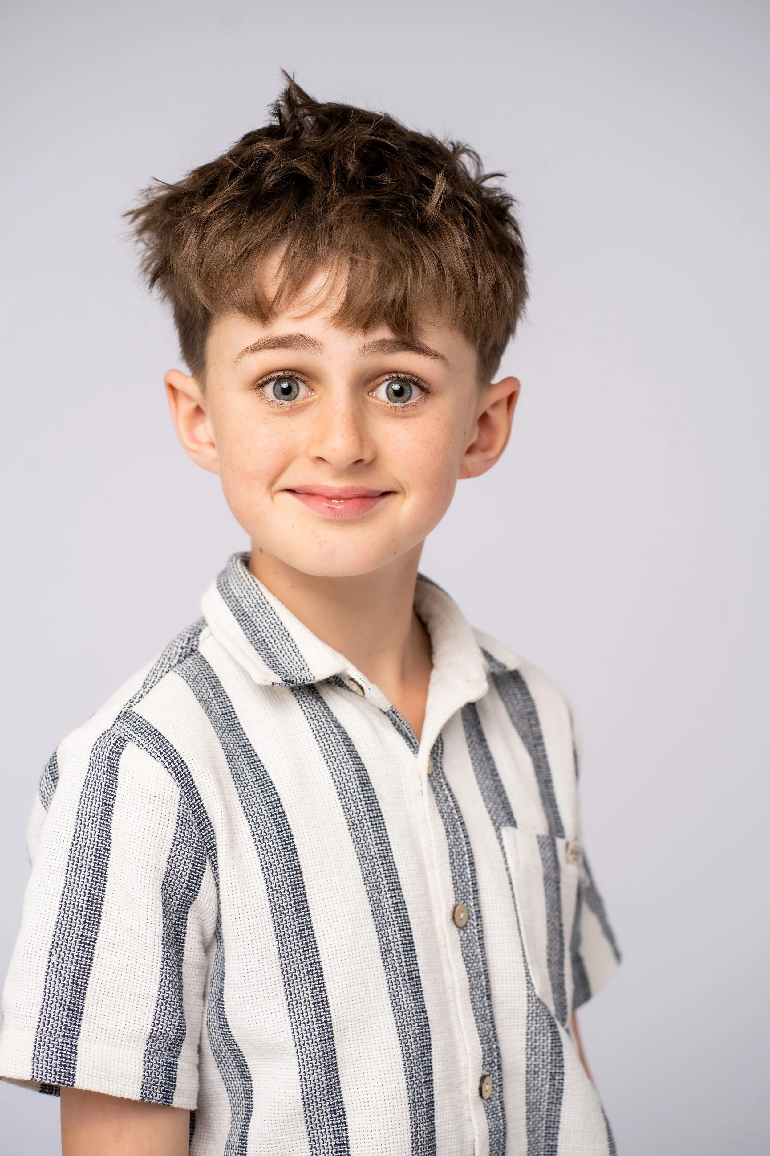A young boy with curly brown hair, blue eyes, and a slight smile, wearing a white and gray striped short-sleeve button-up shirt, standing against a plain light gray background.