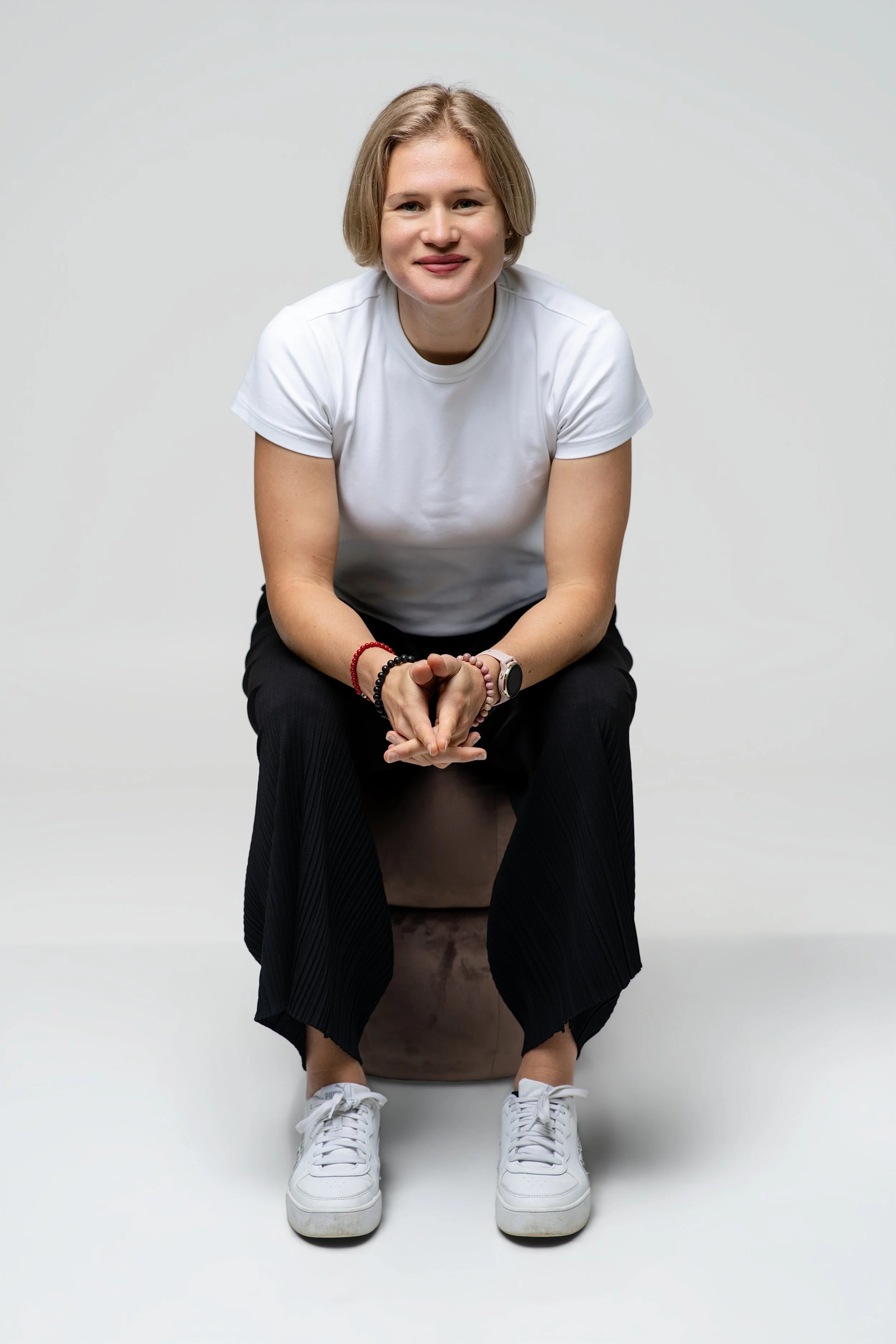 A woman with short blonde hair, wearing a white t-shirt, black pants, and white sneakers, sitting on a brown ottoman against a plain white background.