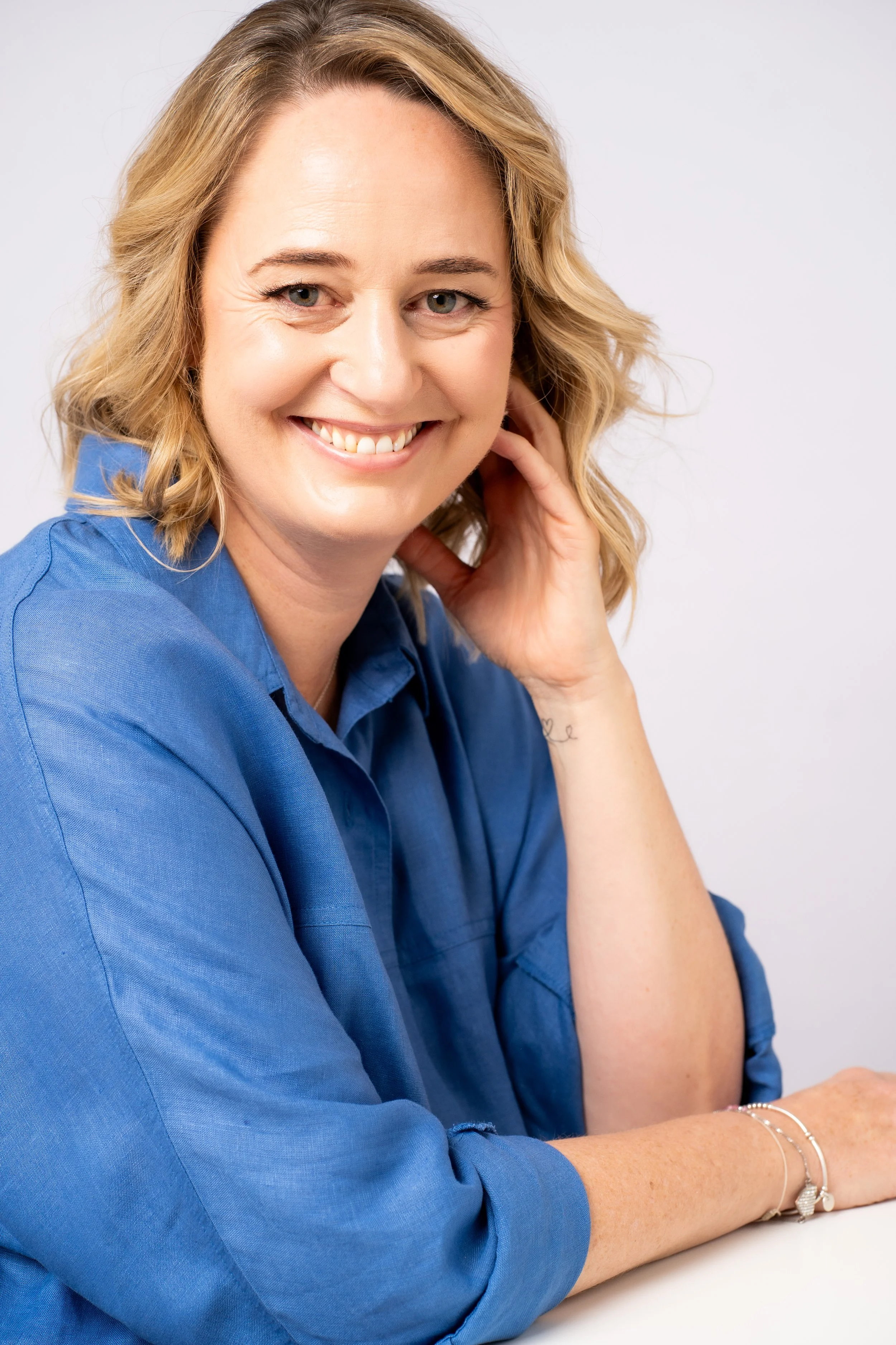 A woman with blonde wavy hair smiling, wearing a blue shirt, and sitting with her hand on her neck against a white background.