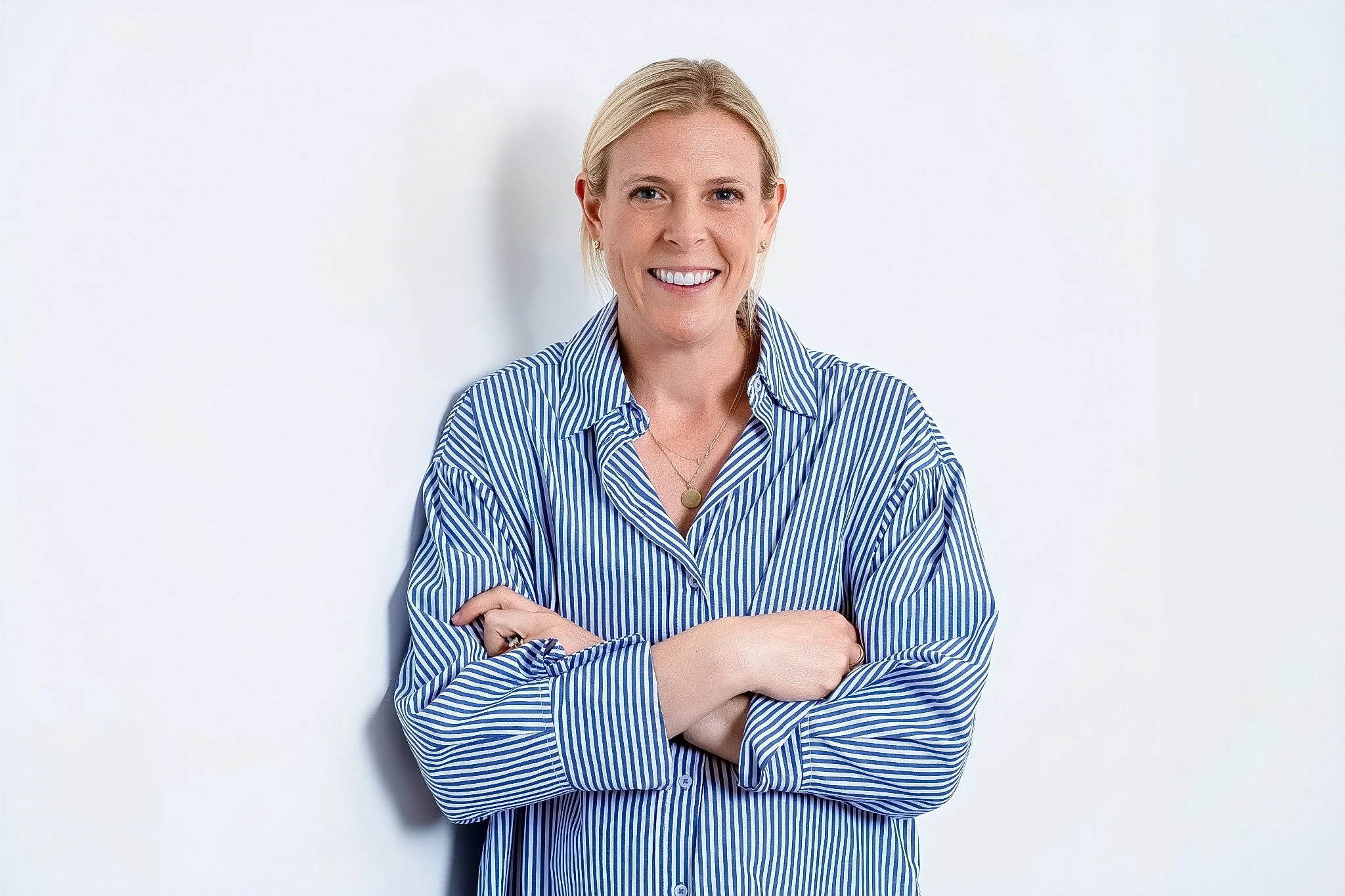 A woman with blonde hair wearing a blue and white striped shirt standing against a plain white background with her arms crossed and smiling.