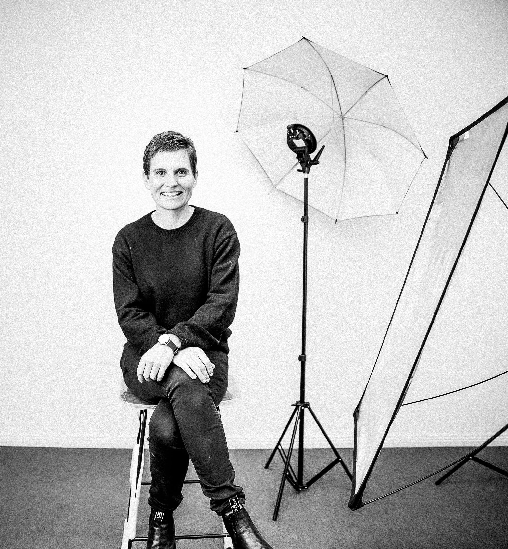 A man with short hair and a wide smile, wearing a black sweater and jeans, sitting on a stool in a photography studio. There is a large umbrella, a light stand, and a diffused light setup around him, indicating a photo shoot environment.