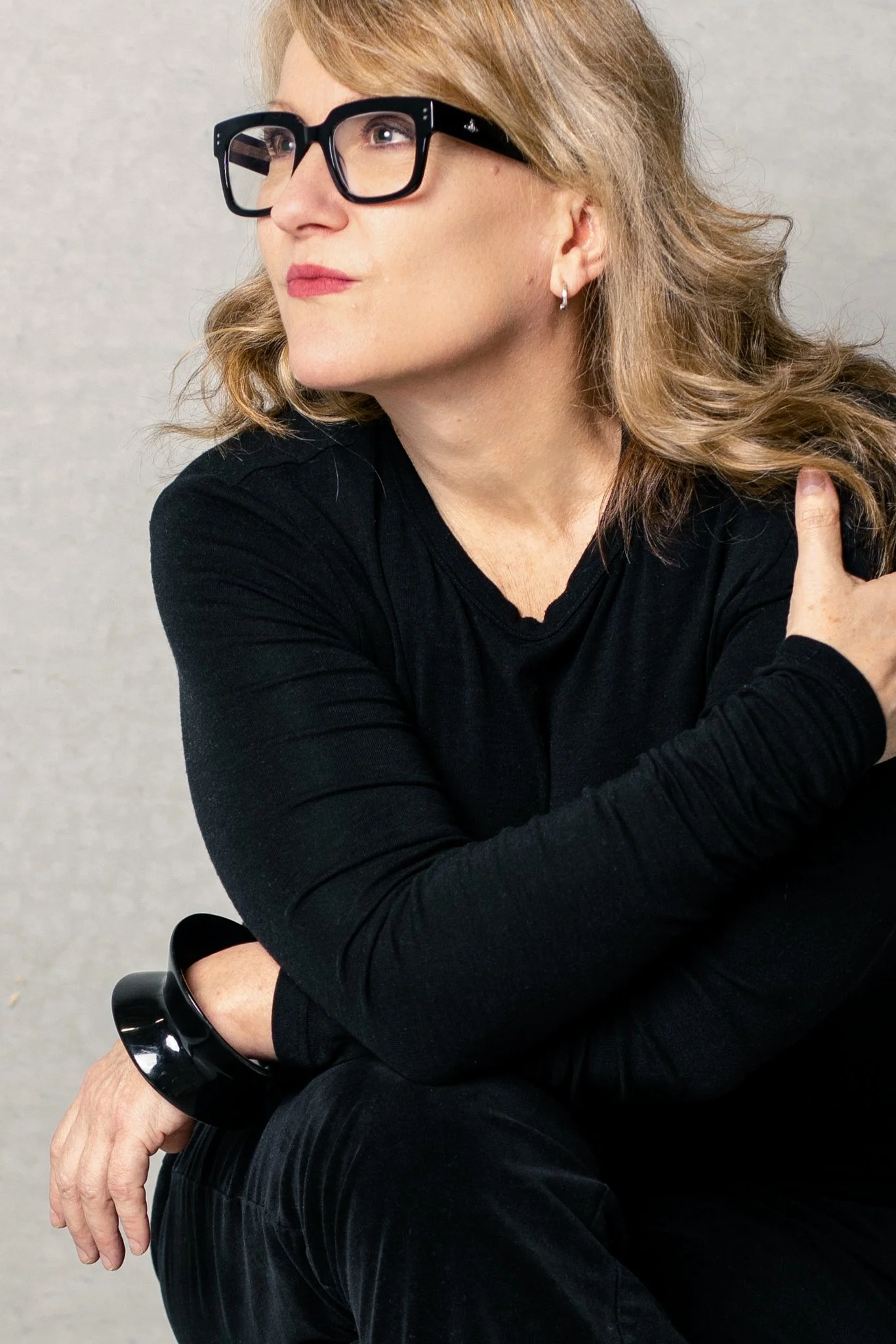 A woman with blonde hair wearing black glasses, a black long sleeve shirt, and a black bracelet, is sitting with her arms crossed and looking to the left against a plain light background.