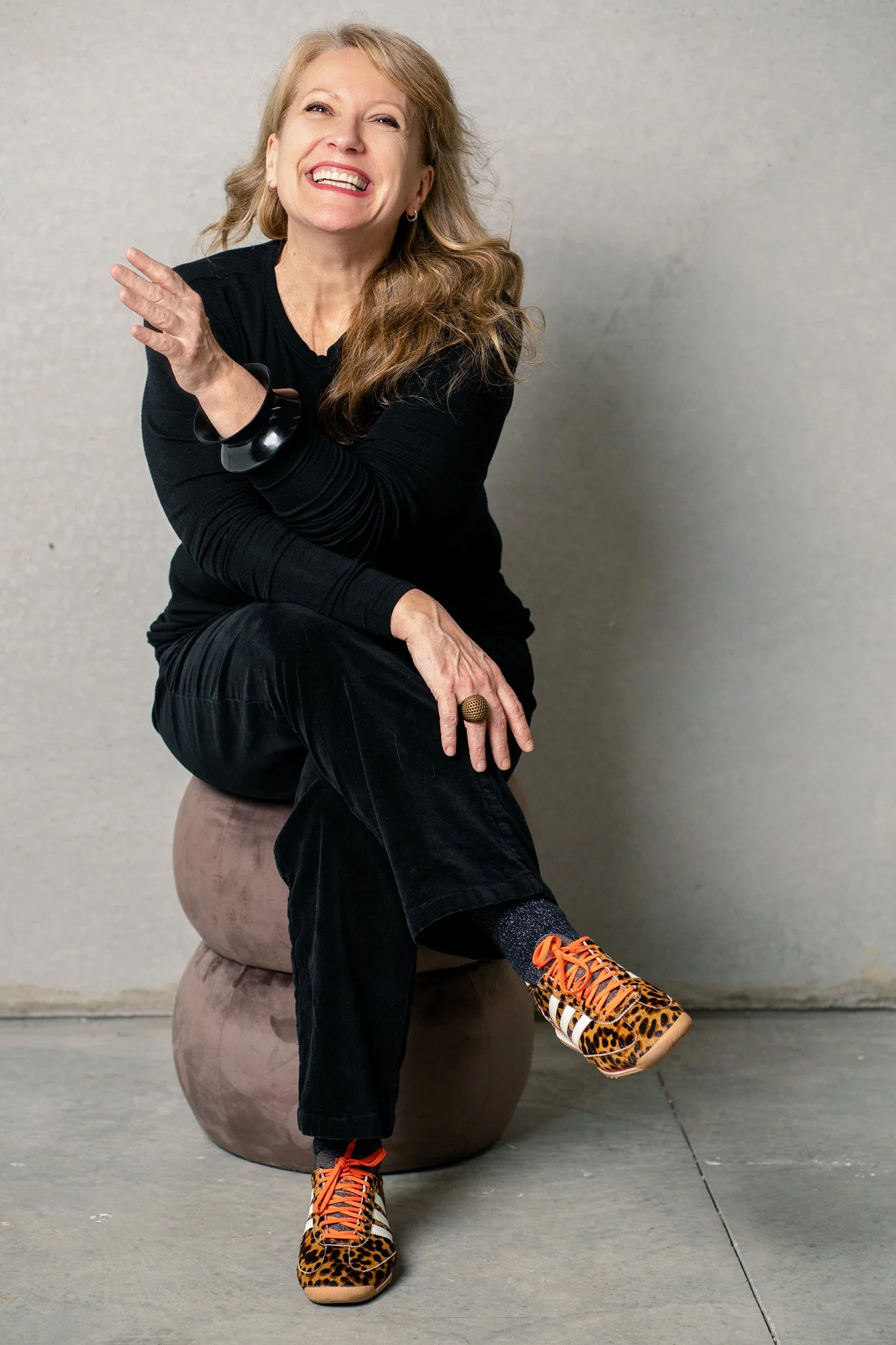 A smiling woman with long light brown hair, wearing a black top, black pants, animal print sneakers with orange laces, and leopard print socks. She is sitting on a decorative stool against a plain wall.