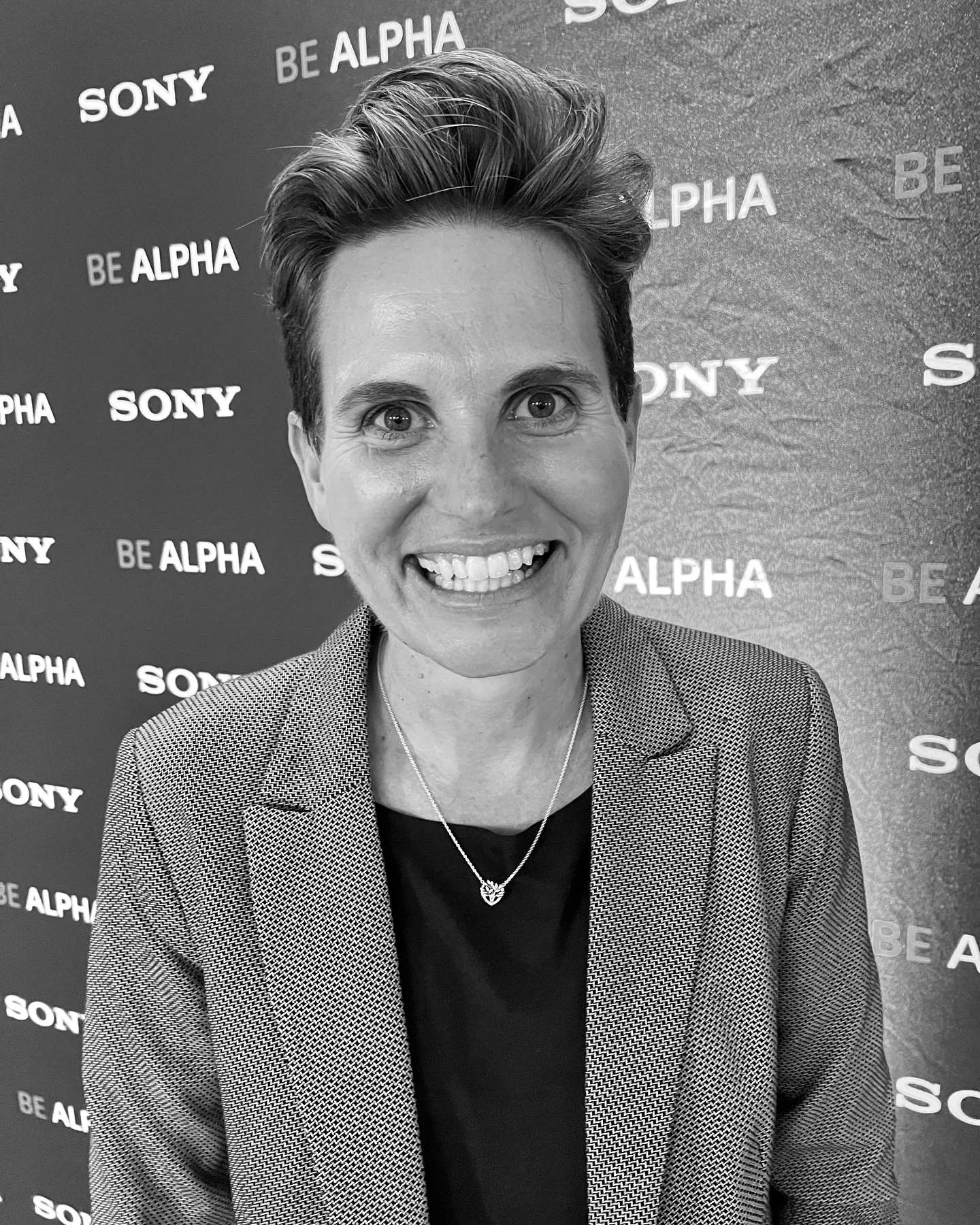 A woman with short hair and a big smile, wearing a blazer, necklace, and black shirt, standing in front of a backdrop with the words 'SONY' and 'BE ALPHA' repeated.
