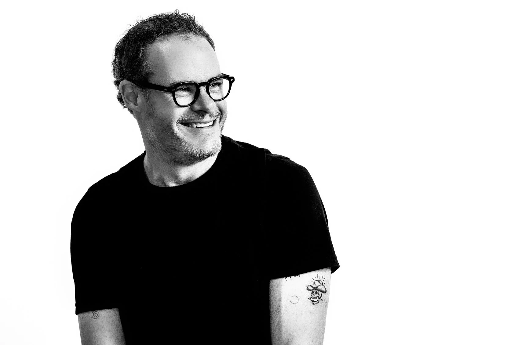 Black and white portrait of a smiling man with glasses, a beard, and a tattoo on his forearm, wearing a black t-shirt, looking to the side against a white background.