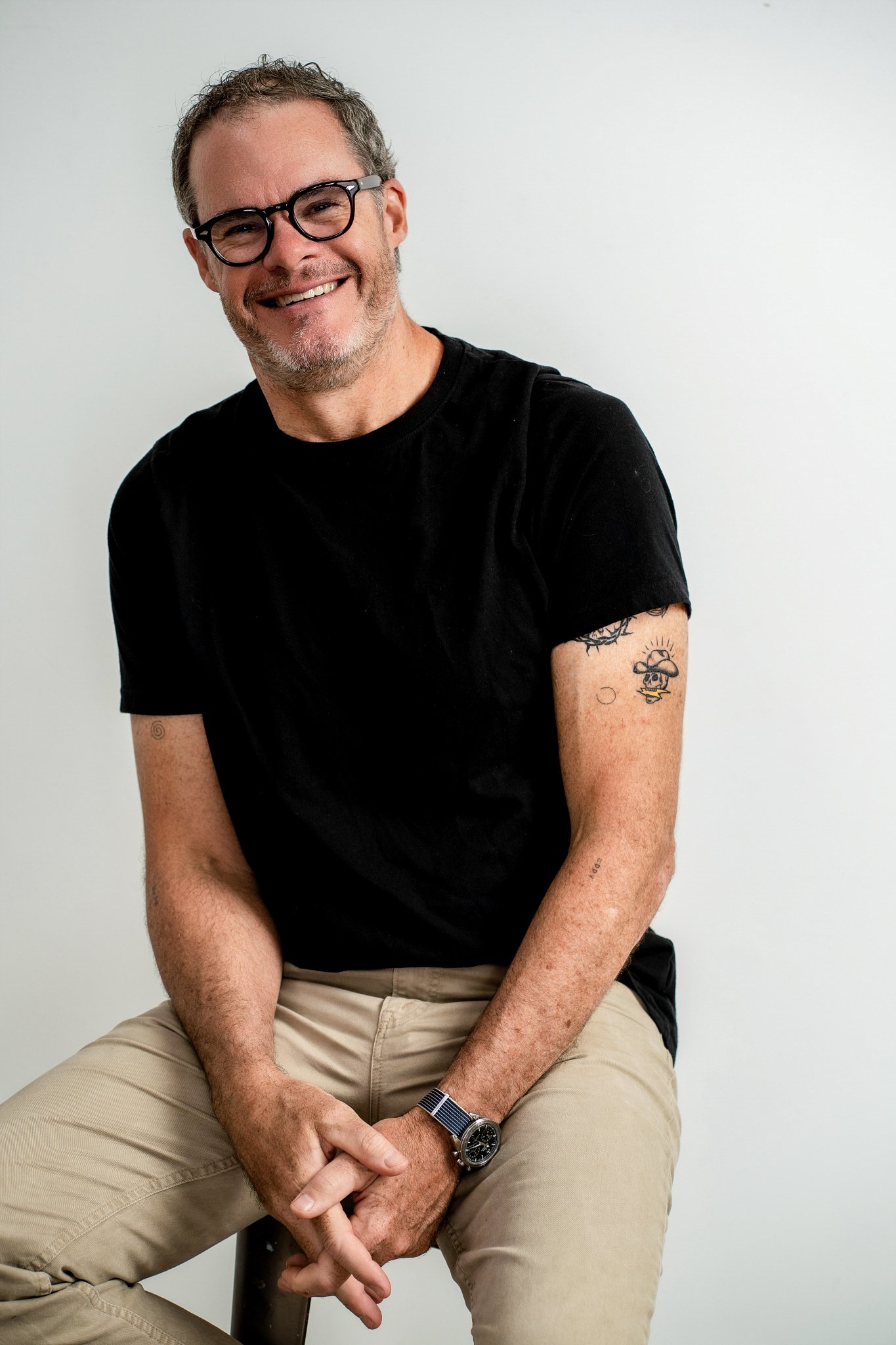 A middle-aged man with glasses, a beard, and tattoos on his arm, smiling while sitting on a chair against a plain white background. He is wearing a black T-shirt, beige pants, a watch, and a bracelet.