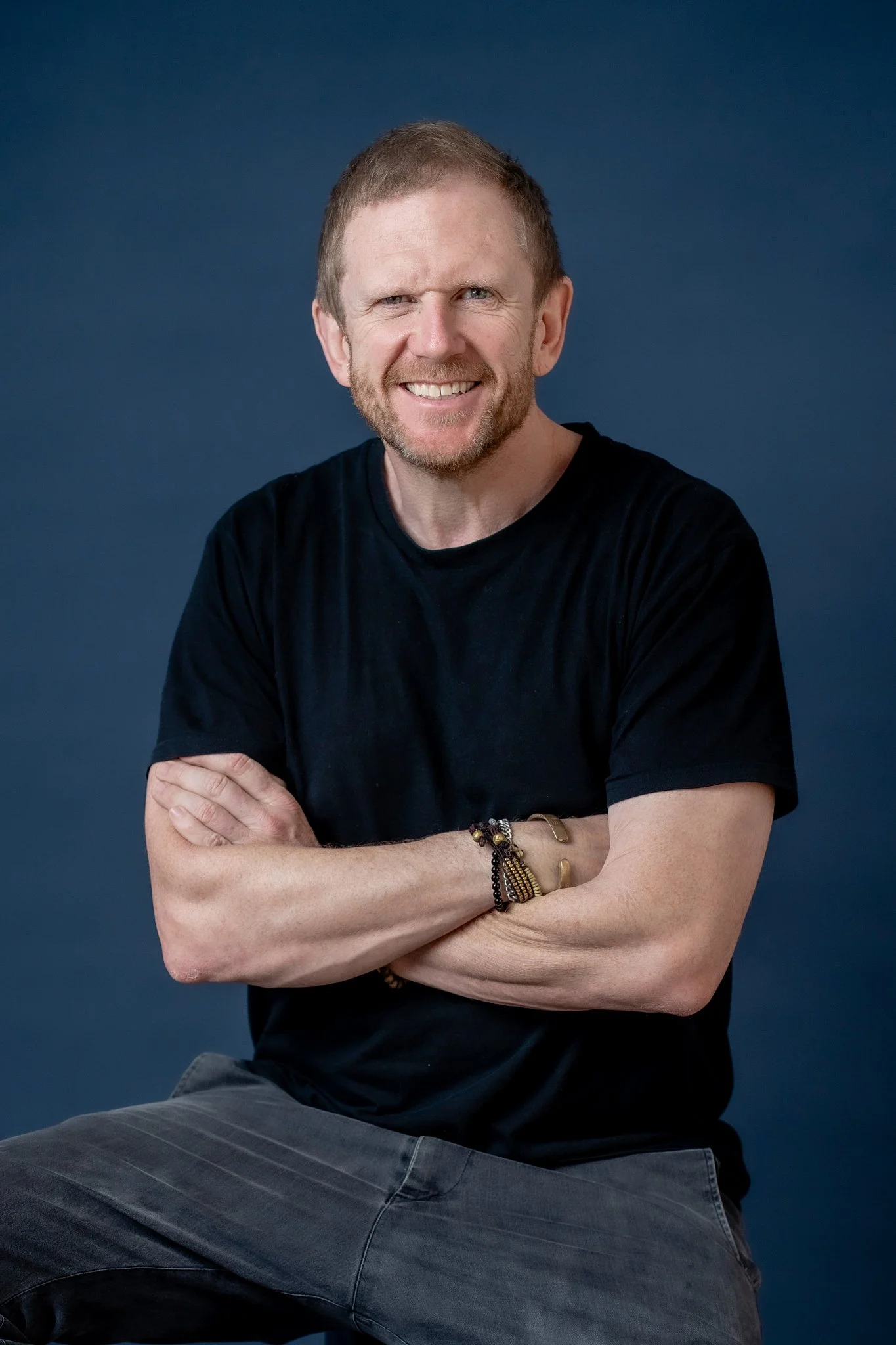 A man with short hair, beard, and blue eyes smiling, with arms crossed, wearing a black t-shirt, bracelet, and rings, sitting against a dark blue background.