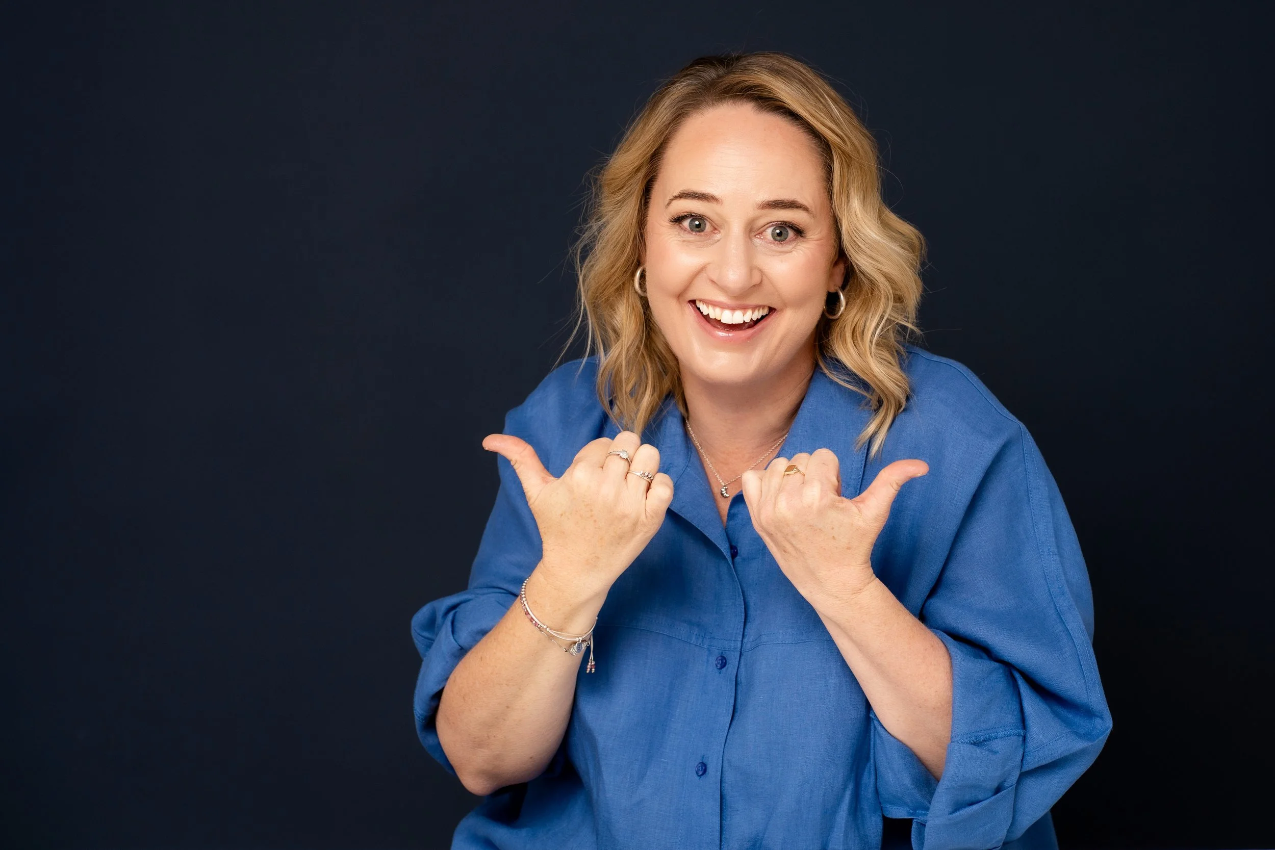 A woman with blonde hair, wearing a blue shirt, smiling broadly, pointing with both hands, against a dark background.