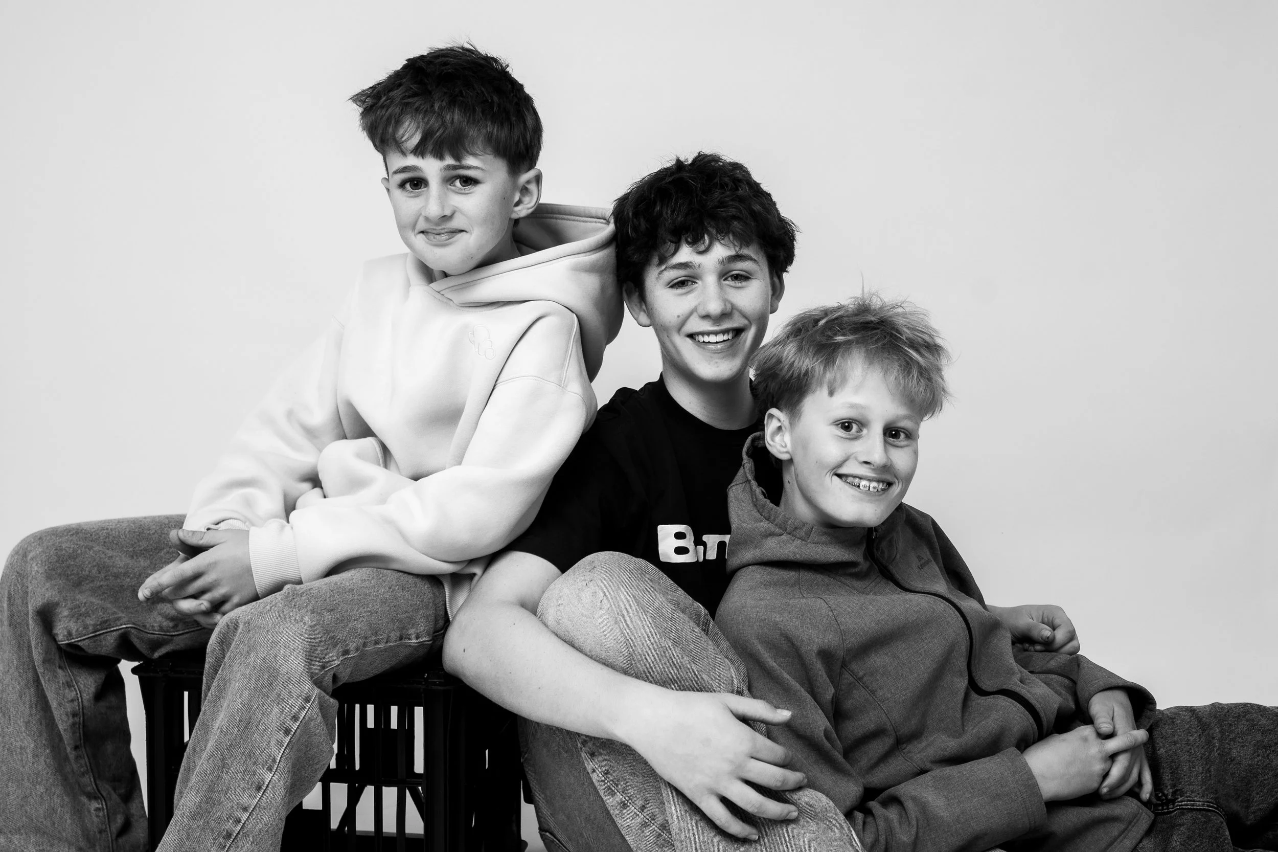 Three boys sitting on a bench posing for a photo in black and white, smiling and looking at the camera.