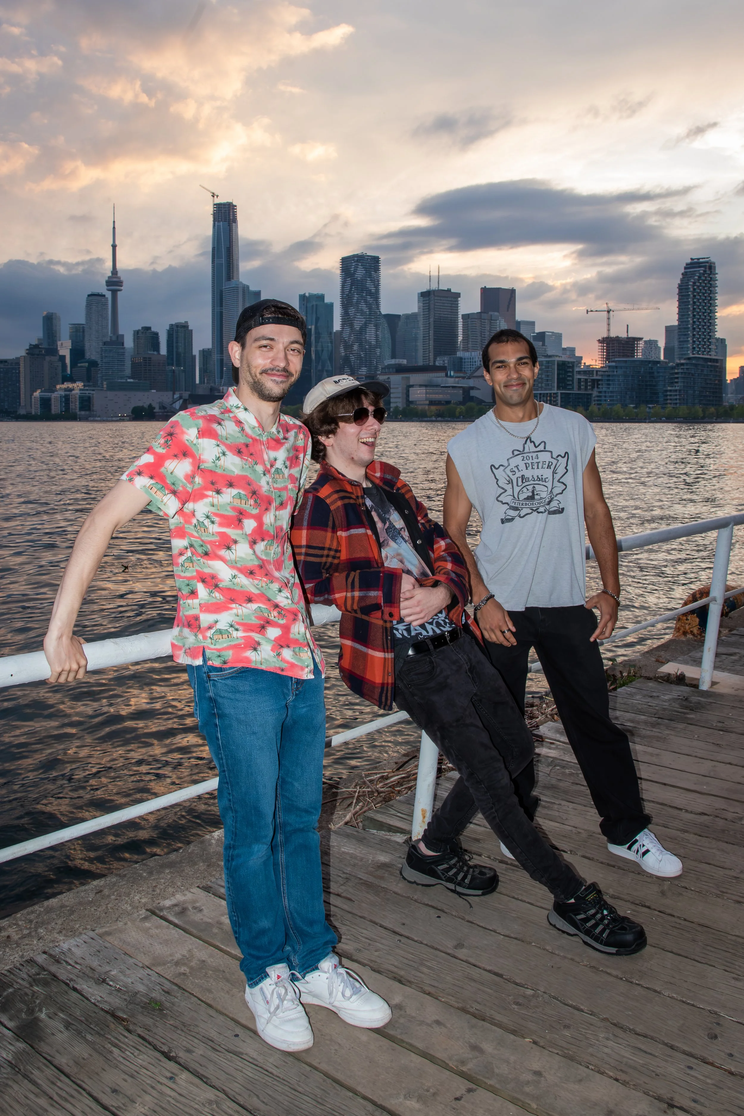Three young men standing together on a wooden dock by water with a city skyline in the background during sunset.